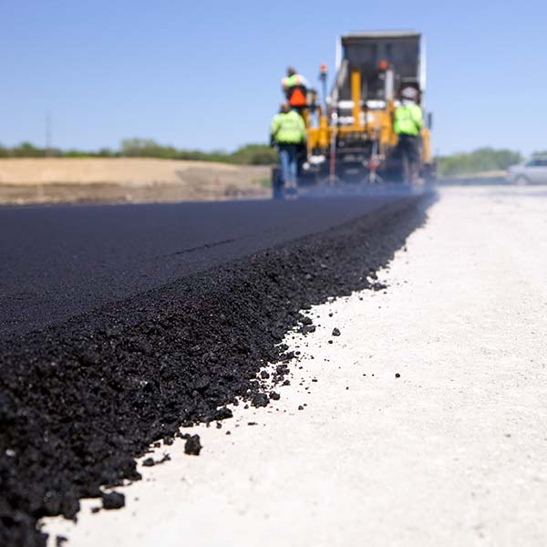 A dump truck is unloading fresh asphalt into a slipform paver, at a new road construction site. Steam is rising from the hot surface directly behind the paver and a car is passing on the cross street. This image could be used equally well for a new driveway project.
