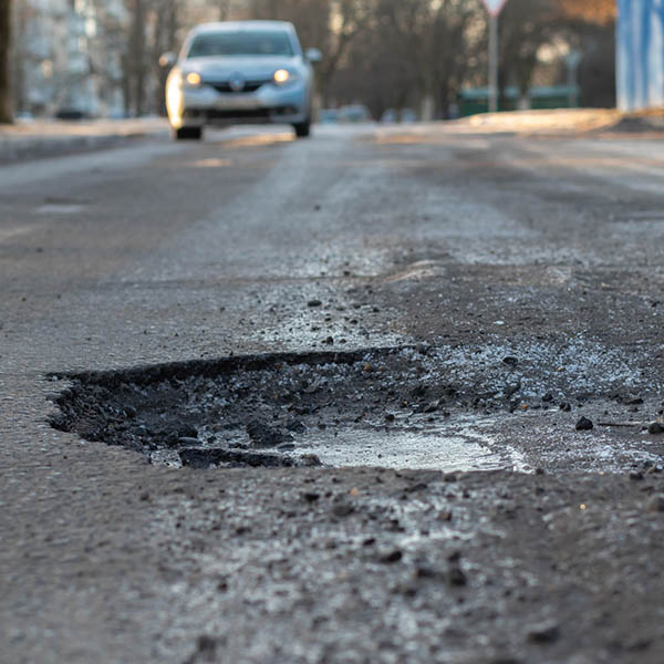 A pothole in the road and a car in the background that is moving towards this pothole