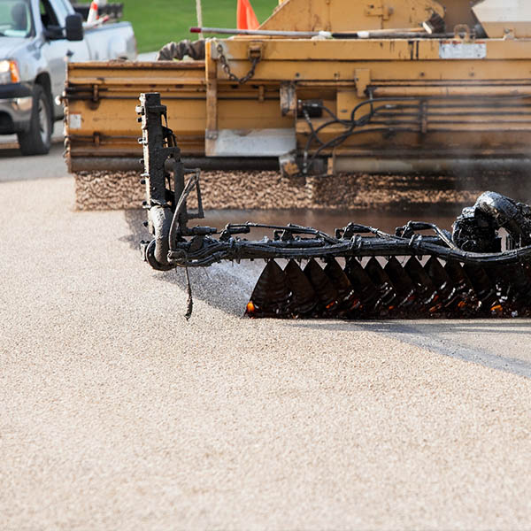 Chip sealing a city street. This image shows the entire chipseal process. The left side has already been reconditioned. On the right a black asphalt seal coat is being applied, behind small chipped rock is falling from an application machine. The rock will be pressed into the asphalt and excess swept up. Chipsealing is a less costly alternative to resurfacing an asphalt road.